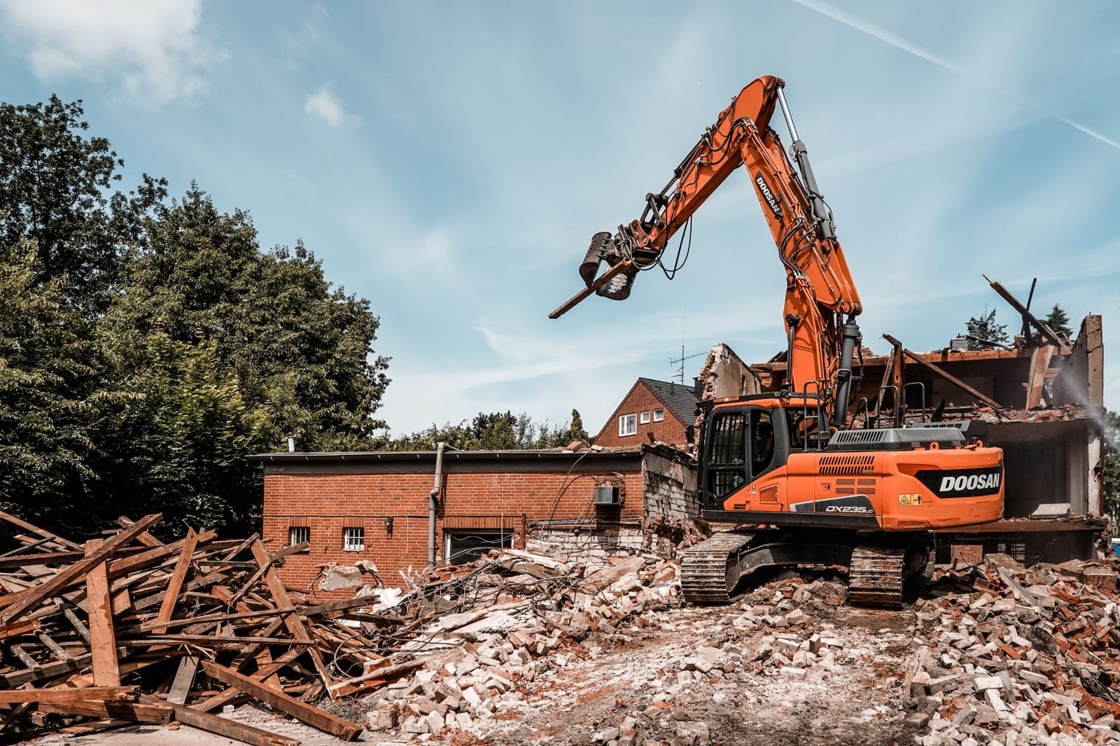 Orangefarbener Abbruchbagger mit Abbruchzange reißt ein Backsteingebäude ab, im Vordergrund liegen sortierbare Trümmer aus Ziegeln und Holzbalken unter blauem Himmel.