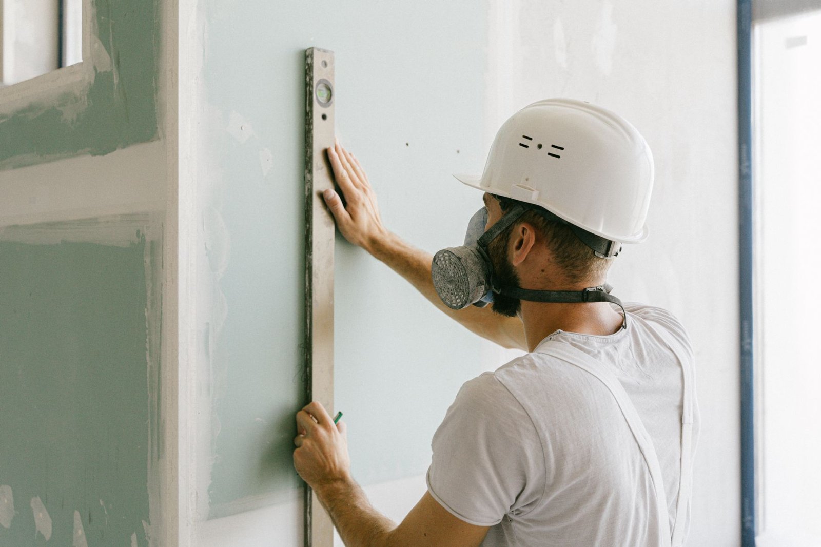 Handwerker mit Schutzhelm und Atemschutzmaske prüft mit einer Wasserwaage die Ausrichtung einer Gipskartonwand im Rohbau – symbolisch für steigende Baupreise im Gewerbeausbau 2026