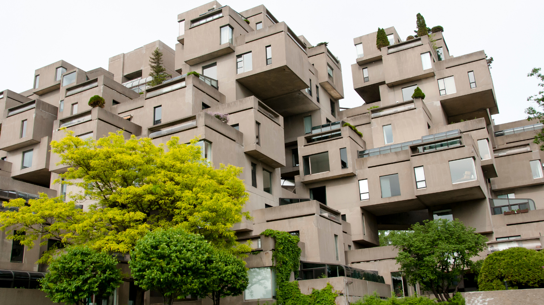 Ein verschachtelter Wohnkomplex mit Wohnungen aus Beton ist vor dem weißen Himmel zu sehen. Pflanzen ringsherum brechen das starre Betonkonzept auf. 